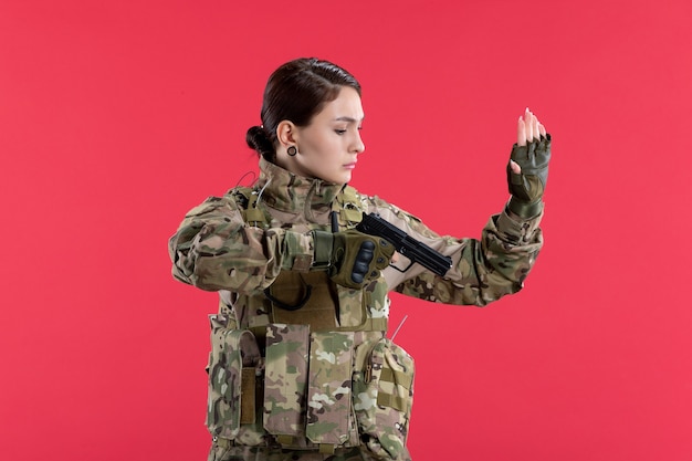 Vue de face d'une femme soldat en tenue de camouflage tenant un pistolet sur un mur rouge