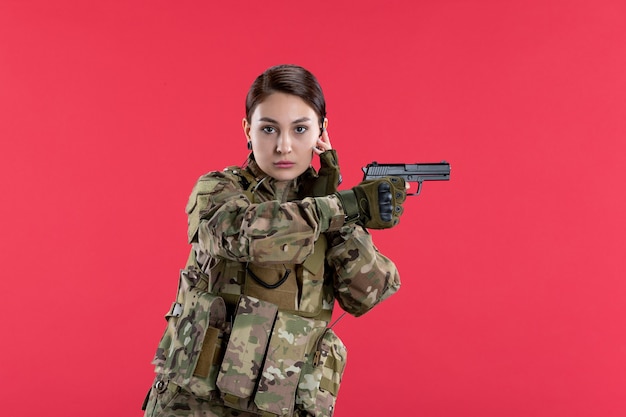 Vue de face d'une femme soldat en tenue de camouflage tenant un pistolet sur un mur rouge