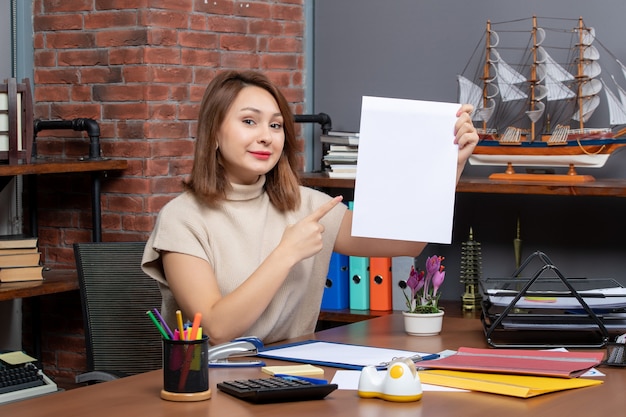 Vue de face femme souriante pointant sur papiers assis au mur