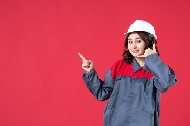 Vue de face d'une femme souriante en uniforme avec un casque et faisant un geste de l'appel vers le haut sur fond rouge isolé