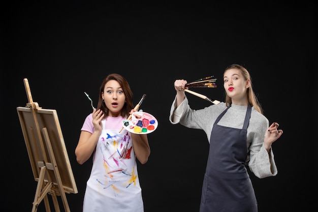 Vue de face femmes peintres tenant des peintures et des glands pour dessiner sur un mur noir photo art artiste couleur travail photo dessiner de la peinture