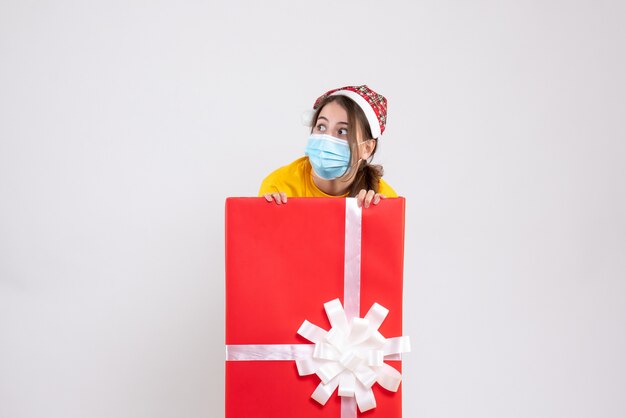 Vue de face fille agitée avec bonnet de Noel debout derrière un grand cadeau de Noël