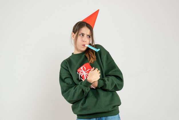 Vue de face fille agitée avec chapeau de fête cachant son cadeau de Noël à l'aide de bruiteur