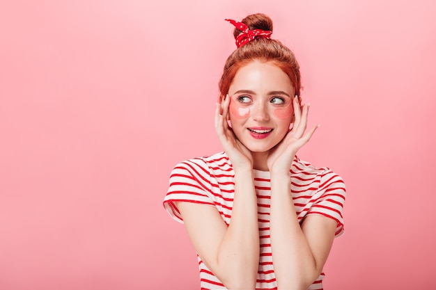 Vue de face d'une fille au gingembre de rêve avec des patchs oculaires. Jolie femme en t-shirt rayé faisant la routine de soins de la peau isolée sur fond rose.