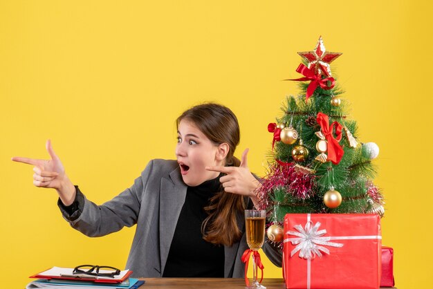 Vue de face fille aux yeux écarquillés assis à la table pointant avec le doigt quelque chose d'arbre de Noël et de cadeaux cocktail