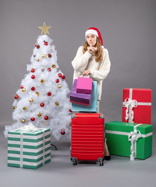 Vue de face fille blonde avec bonnet de noel tenant valise rouge et sacs à provisions montrant signe chut