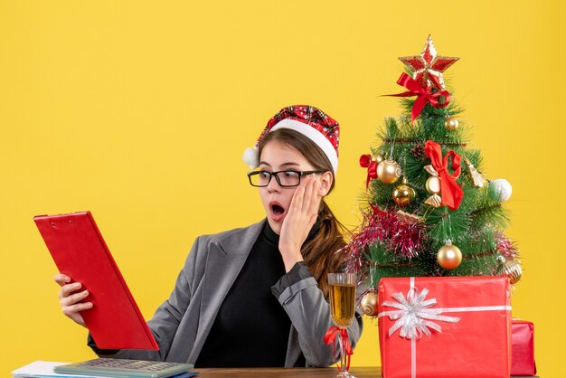 Vue de face fille choquée avec chapeau de Noël assis à la table en regardant le fichier de document arbre de Noël et cadeaux cocktail