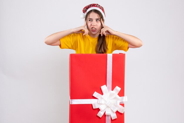 Vue de face fille en colère avec bonnet de Noel debout derrière un gros cadeau de Noël mettant les doigts