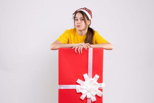 Vue de face fille confuse avec bonnet de Noel debout derrière un grand cadeau de Noël