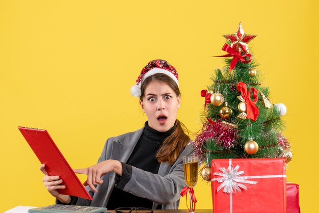 Vue de face fille confuse avec chapeau de Noël assis à la table dossier de document arbre de Noël et cadeaux cocktail sur fond jaune de table