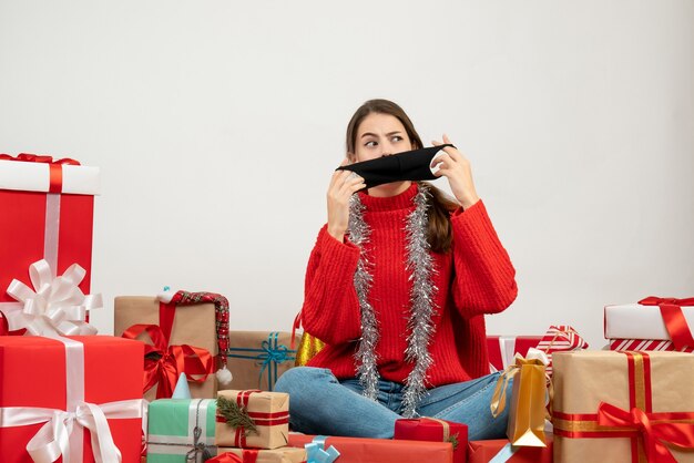 Vue de face fille confuse avec pull rouge décollant son masque assis autour de cadeaux