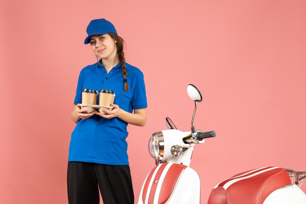 Vue de face d'une fille de courrier heureuse debout à côté d'une moto tenant un café sur fond de couleur pêche pastel