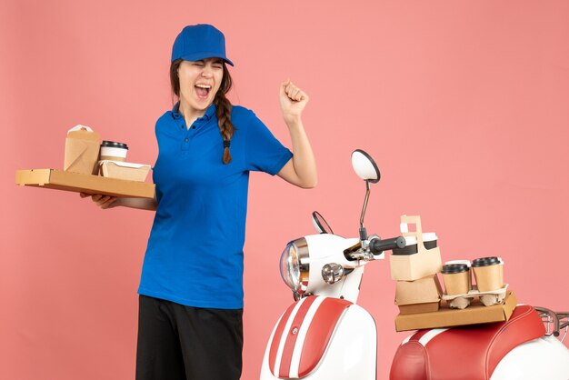 Vue de face d'une fille de coursier fière et émotionnelle debout à côté d'une moto tenant du café et de petits gâteaux sur fond de couleur pêche pastel