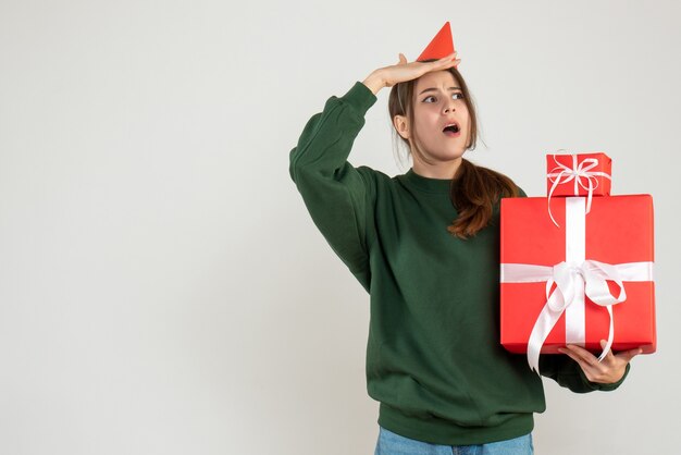 Vue de face fille curieuse avec chapeau de fête observant tenant ses cadeaux de Noël