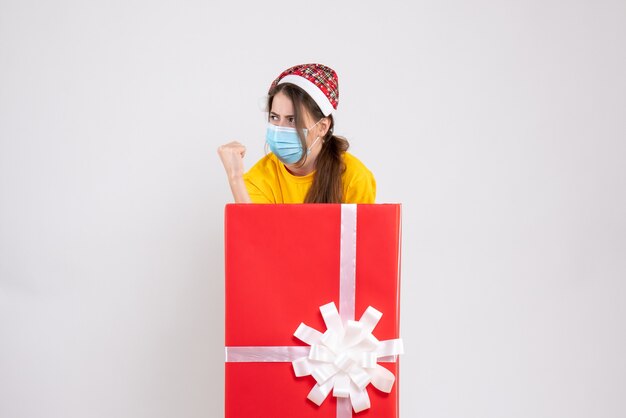 Vue de face fille déterminée avec bonnet de Noel debout derrière un grand cadeau de Noël