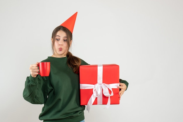 Vue de face fille excitée avec chapeau de fête tenant son cadeau de Noël et une tasse