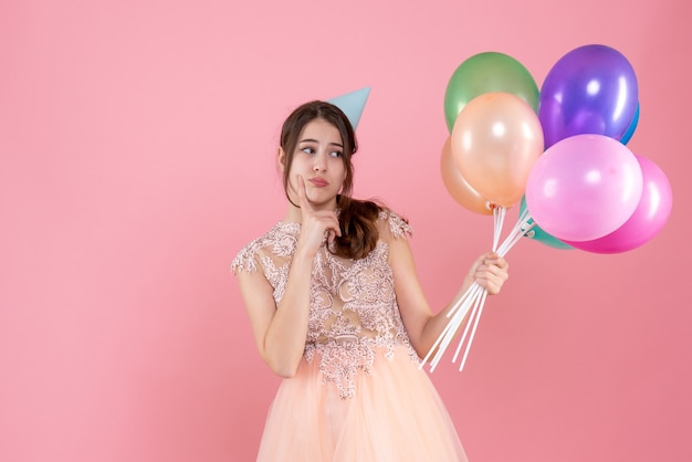 Vue de face fille de fête confuse avec chapeau de fête tenant des ballons