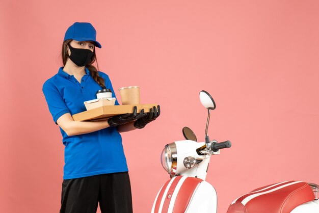 Vue de face d'une fille de messagerie concentrée portant des gants de masque médical debout à côté d'une moto tenant des petits gâteaux de café sur fond de couleur pêche pastel