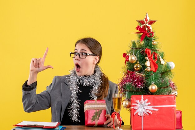 Vue de face fille perplexe avec des lunettes assis à la table pointant avec le doigt jusqu'à l'arbre de Noël et des cadeaux cocktail