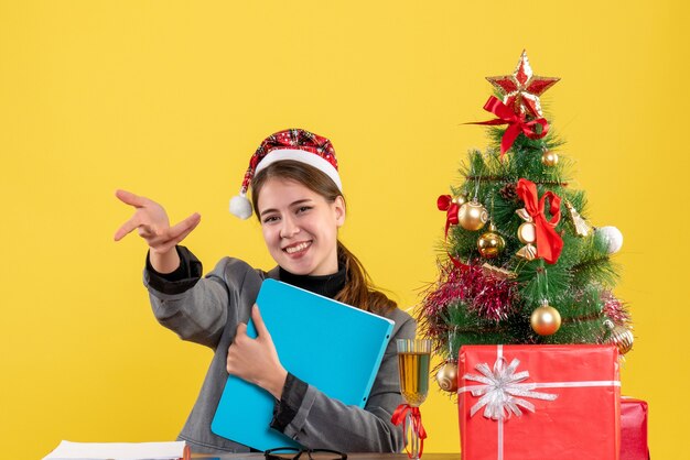 Vue de face fille souri avec chapeau de Noël assis à la table de Noël arbre et cadeaux cocktail