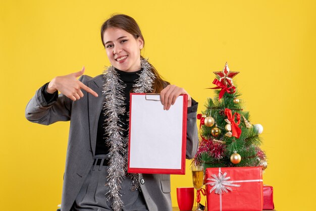 Vue de face fille sourit montrant des documents debout près de l'arbre de Noël et des cadeaux cocktail