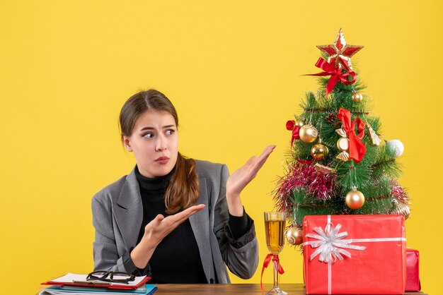 Vue de face fille surprise assise au bureau montrant un arbre de Noël et un cocktail de cadeaux