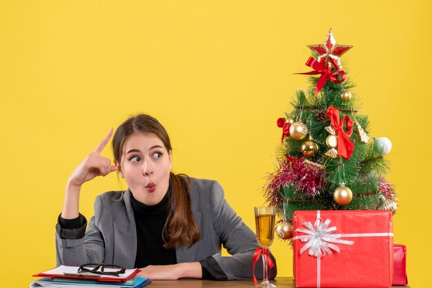 Vue de face fille surprise assise à la table montrant quelque chose d'arbre de Noël et de cadeaux cocktail