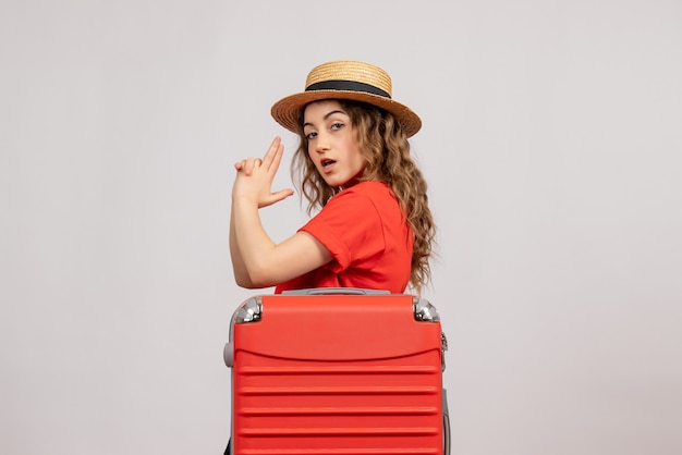 Vue de face de la fille de vacances avec sa valise faisant pistolet à doigt debout sur le mur blanc