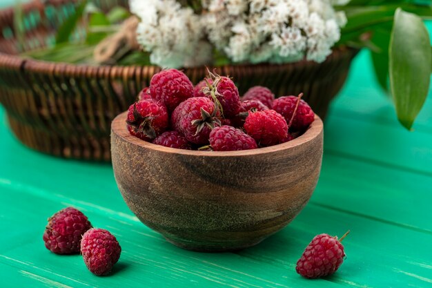 Vue de face de framboises dans un bol en bois avec des fleurs sur une surface verte