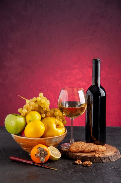 Vue de face fruits frais dans un bol pomme coing raisins kaki bouteille de vin verre de vin cookies sur planche de bois sur table rouge