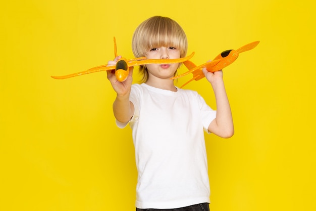 Vue de face garçon blond jouant avec des avions jouets orange en t-shirt blanc sur sol jaune