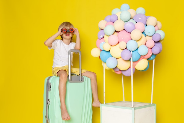 Vue de face garçon blond en t-shirt blanc assis sur le sac vert d'eau avec des ballons à air multicolores sur le sol jaune