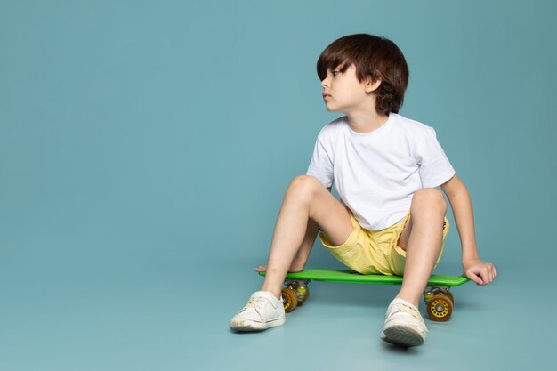 Une vue de face garçon mignon adorable doux mignon en t-shirt blanc équitation planche à roulettes sur l'espace bleu