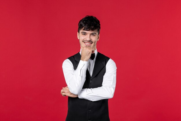 Vue de face d'un garçon souriant et satisfait en uniforme avec un nœud papillon sur le cou sur un mur rouge