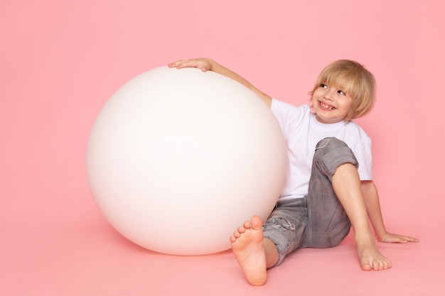 Une vue de face garçon souriant en t-shirt blanc jouant avec une balle ronde blanche sur le sol rose