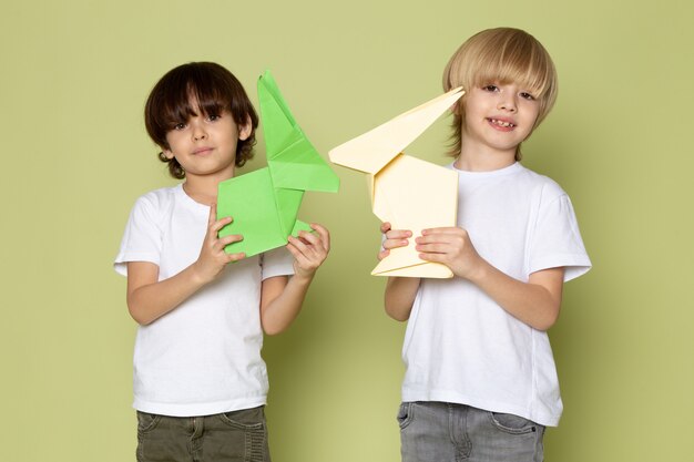 Une vue de face des garçons souriants en t-shirts blancs tenant des jouets en papier sur le mur de couleur pierre