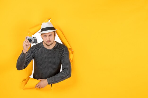 Vue de face d'un gars concentré avec un chapeau tenant une caméra dans un mur jaune déchiré