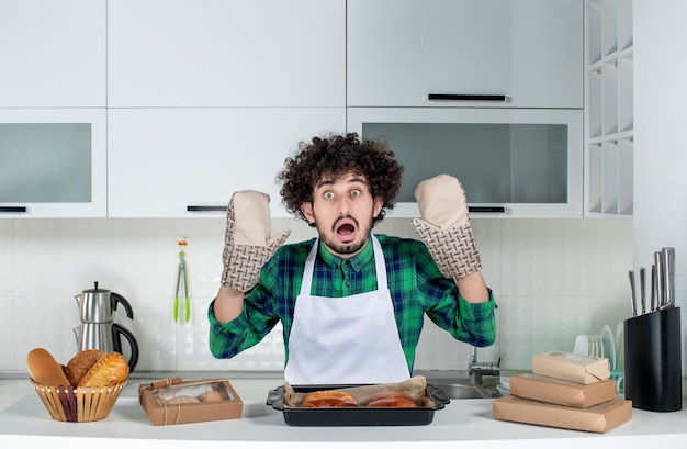 Photo gratuite vue de face d'un gars effrayé portant un support debout derrière une table avec une pâtisserie fraîchement préparée dans la cuisine blanche
