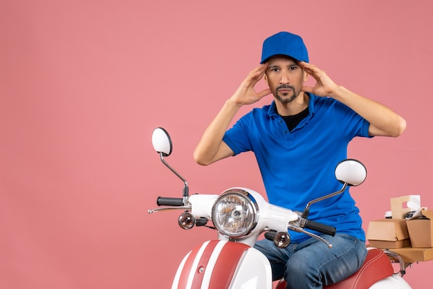 Photo gratuite vue de face d'un gars de messagerie concentré portant un chapeau assis sur un scooter sur fond de pêche pastel