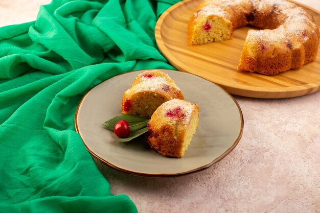 Une vue de face gâteau aux fruits cuits au four de délicieuses tranches avec des cerises rouges à l'intérieur et du sucre en poudre à l'intérieur de la plaque grise ronde sur rose