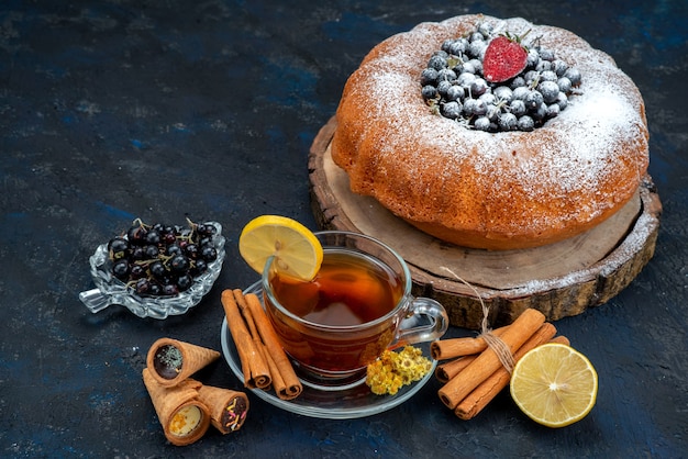 Une vue de face gâteau aux fruits délicieux et rond formé avec du bleu frais, des baies et avec une tasse de thé sur noir, gâteau biscuit sucre sucré