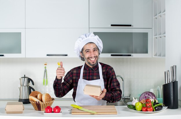 Vue de face happy male chef holding box abandonnant le pouce dans la cuisine