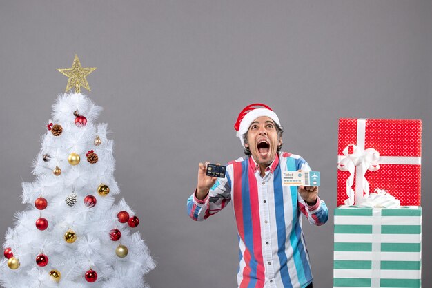 Vue de face happy man holding card et billet de voyage autour de l'arbre de Noël et des cadeaux