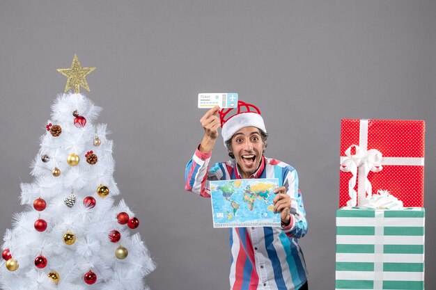 Vue de face heureux jeune homme avec bonnet de noel printemps spirale tenant carte du monde et billet de voyage