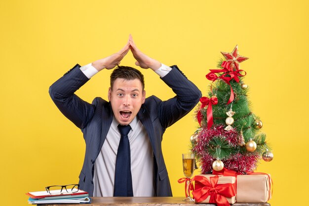 Vue de face de l'heureux jeune homme faisant maison de toit avec ses mains assis à la table près de l'arbre de Noël et des cadeaux sur jaune