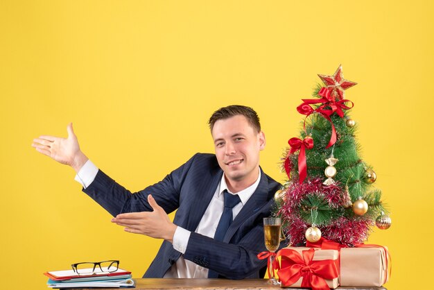 Vue de face de l'heureux jeune homme montrant quelque chose assis à la table près de l'arbre de Noël et des cadeaux sur jaune