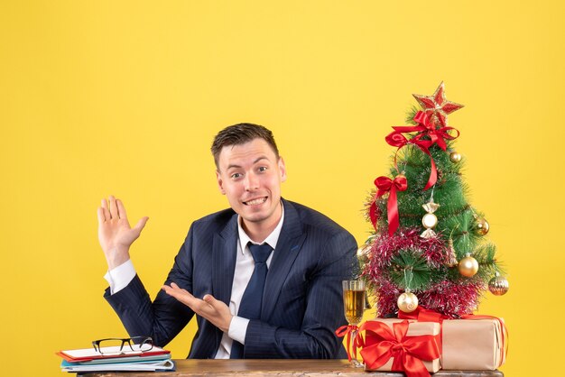 Vue de face de l'heureux jeune homme pointant vers. fond assis à la table près de l'arbre de Noël et des cadeaux sur jaune