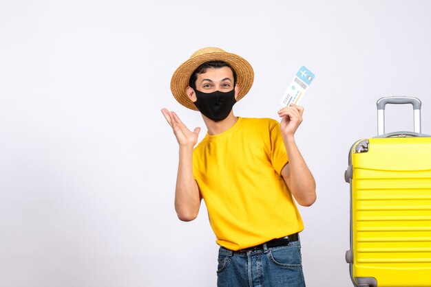 Vue de face heureux jeune homme en t-shirt jaune debout près de valise jaune tenant un billet de voyage