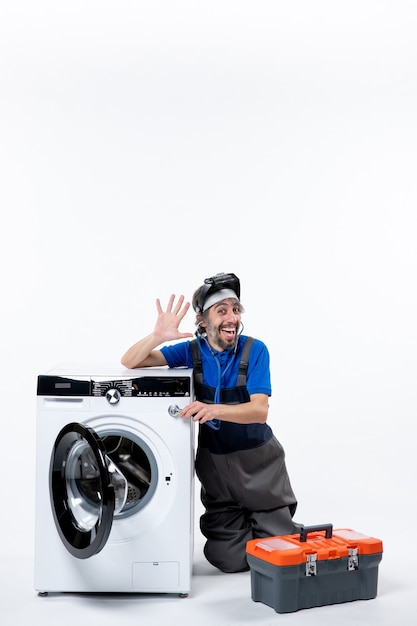 Vue de face heureux réparateur assis près d'une machine à laver hélant quelqu'un sur un espace blanc