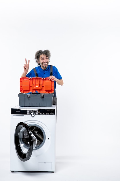 Vue de face heureux réparateur faisant le signe de la victoire derrière la machine à laver sur un espace blanc